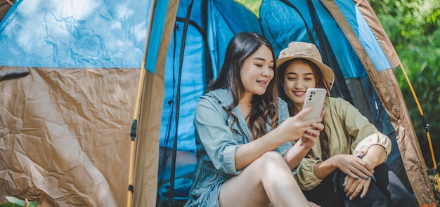 正面図テントの前に座っている若いアジアのきれいな女性と彼女のガールフレンドは携帯電話を使用して一緒に幸せで森のキャンプ中に写真を撮ります