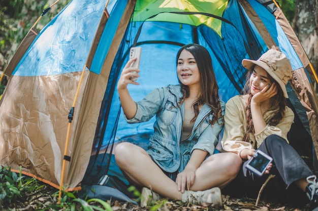 Front view Young Asian pretty woman and her girlfriend sitting at front of tent use mobile phone take photo during camping in forest with happiness together