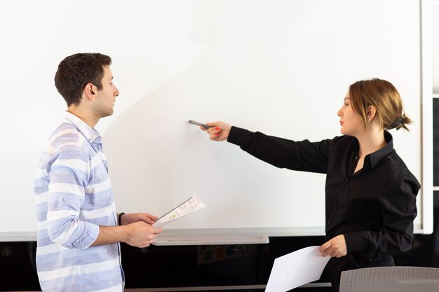 A front view young attractive businesswoman in black shirt along with young man discussing graphics on the desk while young lady presents her work reading document job building presentation