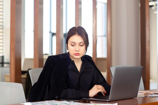 Вид спереди молодой привлекательной бизнес-леди в черной рубашке черный пиджак, используя свой серебряный ноутбук, работающих внутри ее офисной работы