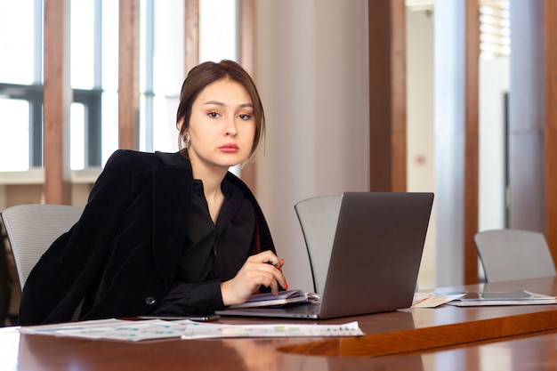 正面の若い魅力的な女性実業家彼女の銀色のラップトップを使用して彼女のオフィスの仕事の仕事の建物の中で作業している黒いシャツの黒いジャケット