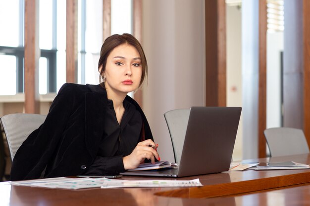A front view young attractive businesswoman in black shirt black jacket using her silver laptop working inside her office work job building