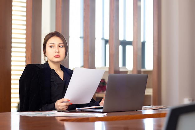 Вид спереди молодой привлекательной бизнес-леди в черной рубашке черный пиджак, используя ее серебристый ноутбук, написание чтения, работающих в ее офисе работа работа здания