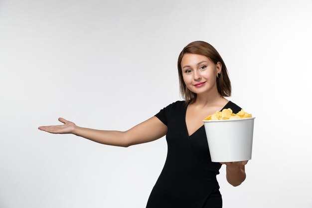 Вид спереди молодая привлекательная женщина в черной рубашке, держащая картофельные чипсы и улыбающаяся на белой поверхности