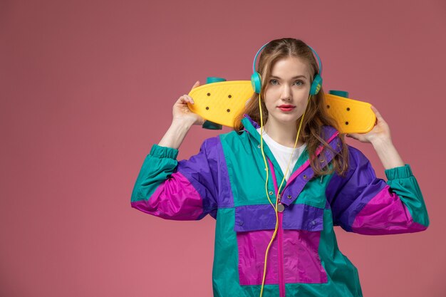 ピンクの壁のモデルの若い女性にスケートボードを保持している音楽を聴いている色のコートで若い魅力的な女性の正面図