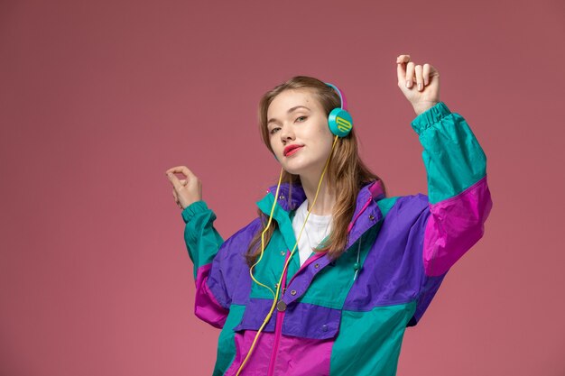 Front view young attractive female in colorful coat listening to music and dancing on pink wall model color female young girl