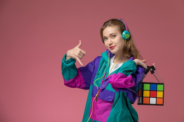 Front view young attractive female in colorful coat smiling listening to music on pink desk model color female young girl