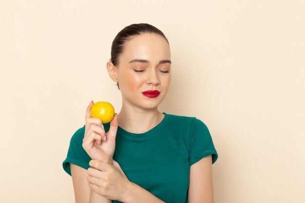 Front view young attractive female in dark green shirt and blue jeans holding yellow lemon on beige