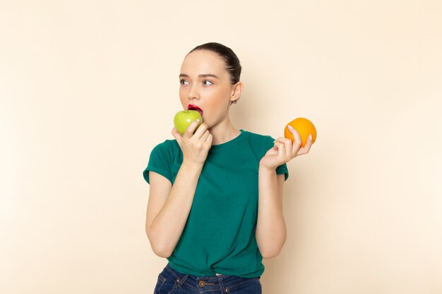 Front view young attractive female in dark green shirt holding orange and apple eating on beige
