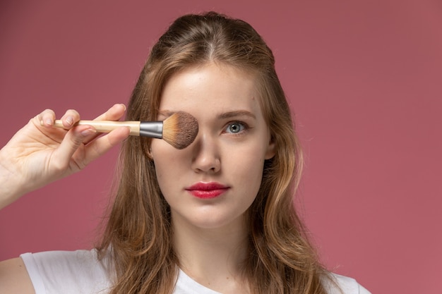 Front view young attractive female doing make-up holding brush on the pink wall model color female young