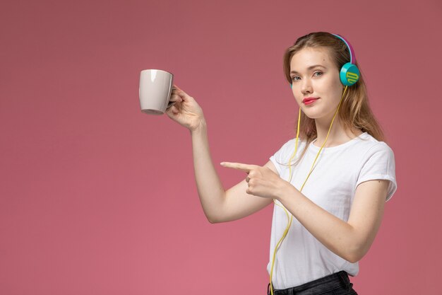 Вид спереди молодая привлекательная женщина, слушающая музыку, держа чашку на розовой стене модель цветная женщина молодая девушка