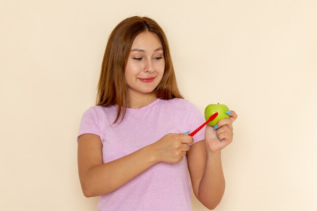 Вид спереди молодая привлекательная женщина в розовой футболке и синих джинсах чистит зеленое яблоко зубной щеткой
