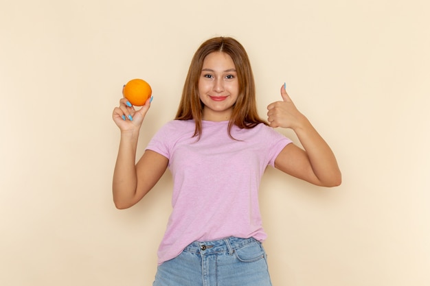 Free photo front view young attractive female in pink t-shirt and blue jeans holding orange and showing awesome sign