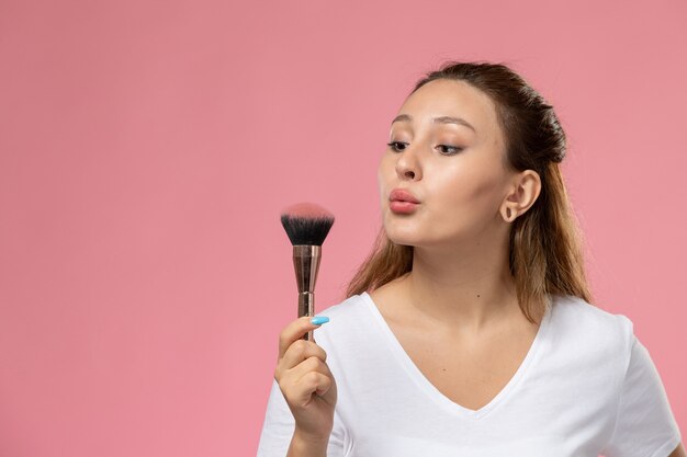 Front view young attractive female in white t-shirt blowing off make-up brush on the pink background