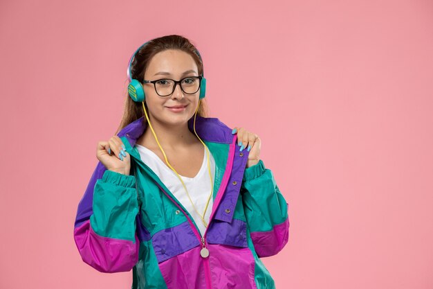 Front view young attractive female in white t-shirt and colored coat listening to music on the pink background