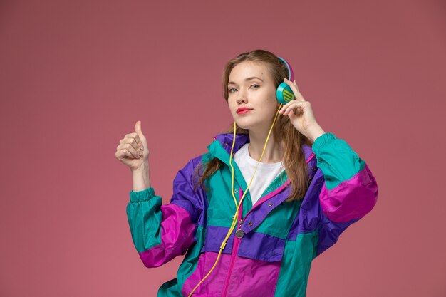 Вид спереди молодая привлекательная женщина в белой футболке, цветное пальто, слушает музыку на розовой стене, цвет модели, девушка, молодая