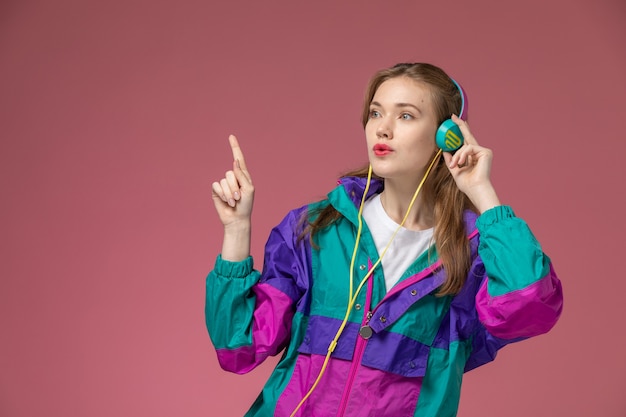 ピンクの壁のモデルの女性のポーズの色の女性の若いで音楽を聴いている白いTシャツ色のコートの正面図若い魅力的な女性