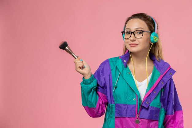 ピンクの背景のポーズモデルの美しさのイヤホンで音楽を聴く白いtシャツ色のコートで正面の若い魅力的な女性