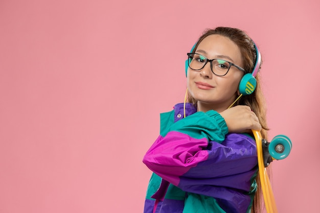 Front view young attractive female in white t-shirt colored coat smiling and holding skateboard on the pink background