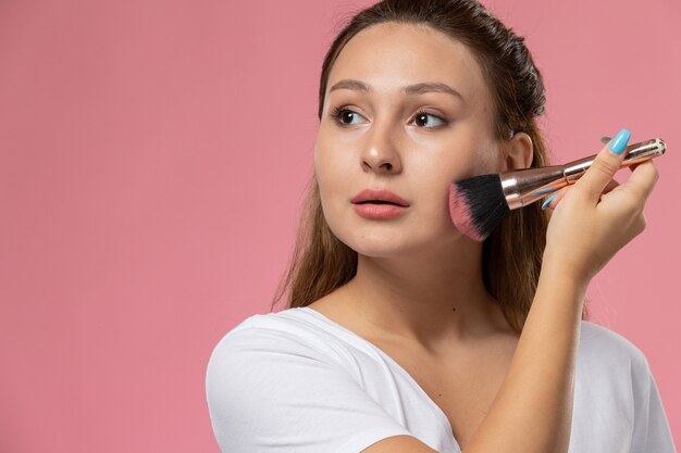 Front view young attractive female in white t-shirt doing make-up on the pink background