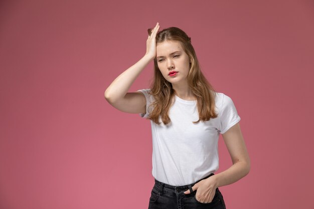 ピンクの壁のモデルの女性のポーズのカラー写真に激しい頭痛を持っている白いTシャツの正面図若い魅力的な女性
