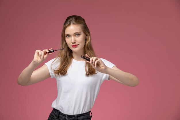 ピンクの机の上の目のマスカラを保持している白いTシャツの正面図若い魅力的な女性モデル色女性の若い女の子