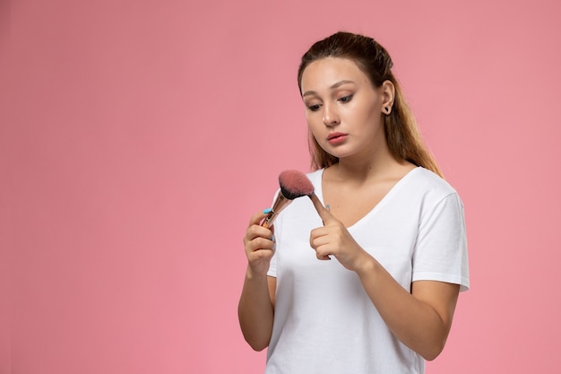 Front view young attractive female in white t-shirt holding make-up brush on the pink background