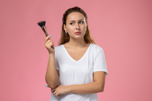 Front view young attractive female in white t-shirt holding make-up brush on the pink background