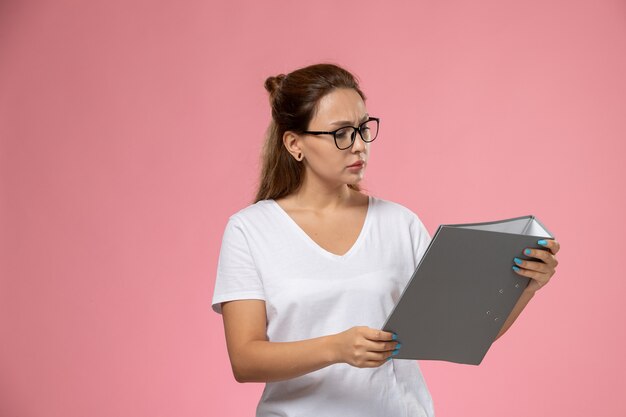 Front view young attractive female in white t-shirt holding and reading grey file on the pink background