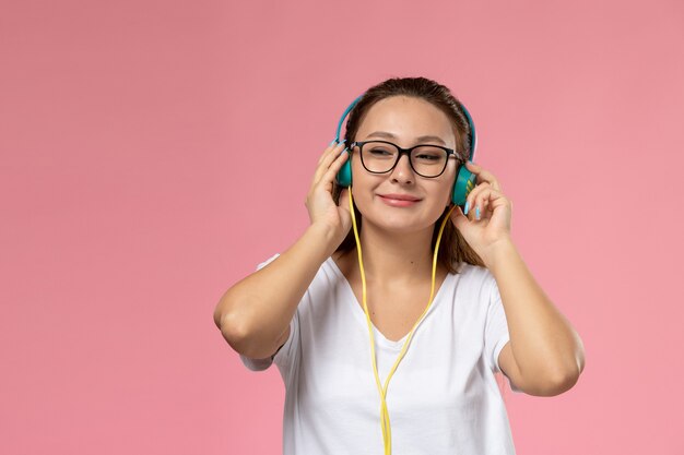 Вид спереди молодая привлекательная женщина в белой футболке, просто позирует и слушает музыку через наушники на розовом фоне