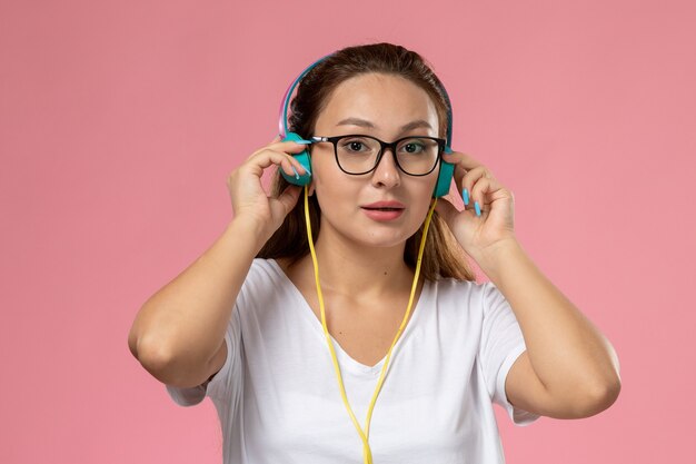 Front view young attractive female in white t-shirt just posing and listening to music via earphones on the pink background