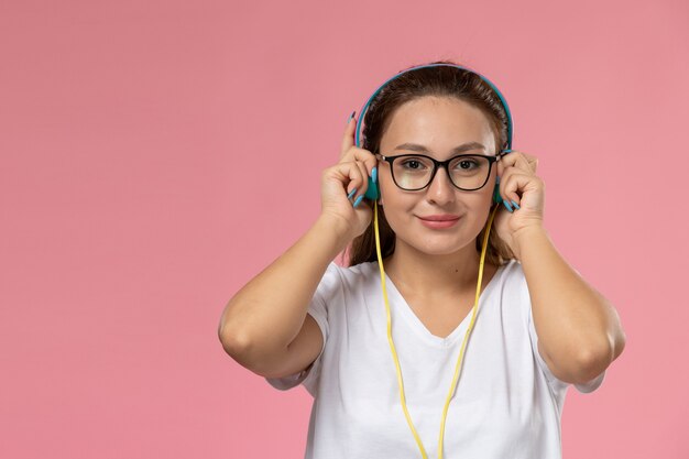 Вид спереди молодая привлекательная женщина в белой футболке, просто позирует и слушает музыку через наушники, улыбаясь на розовом фоне