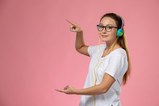 ピンクの背景にわずかな笑顔でポーズをとって音楽を聴く白いtシャツの正面の若い魅力的な女性