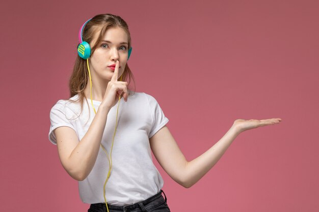 핑크 책상 모델 여성 포즈 색상에 침묵 기호를 보여주는 이어폰을 통해 음악을 듣고 흰색 티셔츠에 전면보기 젊은 매력적인 여성 photo
