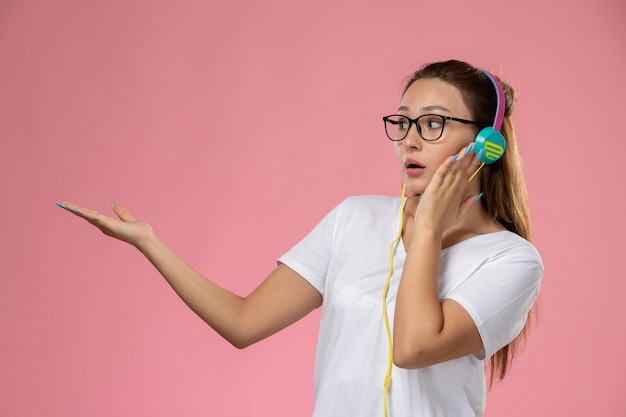 Front view young attractive female in white t-shirt listening to music with earphones on the pink background