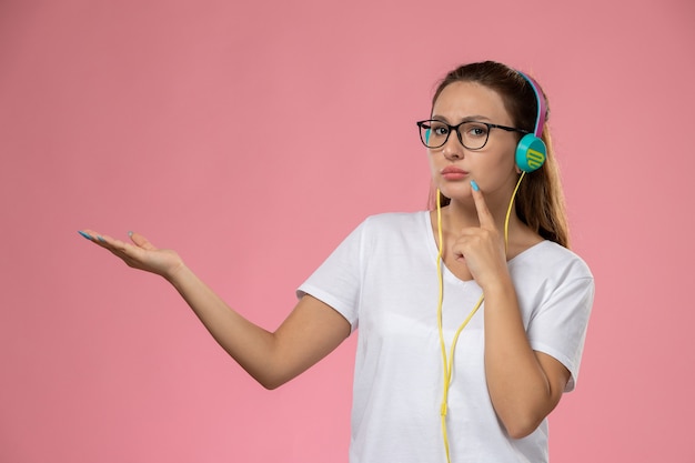 Front view young attractive female in white t-shirt listening to music with headphones posing on the pink desk