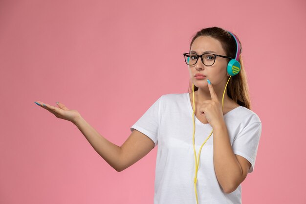 ピンクの机の上でポーズをとってヘッドフォンで音楽を聴く白いtシャツの正面の若い魅力的な女性