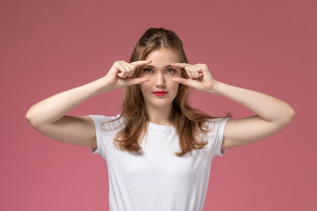 Front view young attractive female in white t-shirt posing and distinguishing her eyes on the pink wall model female pose color photo