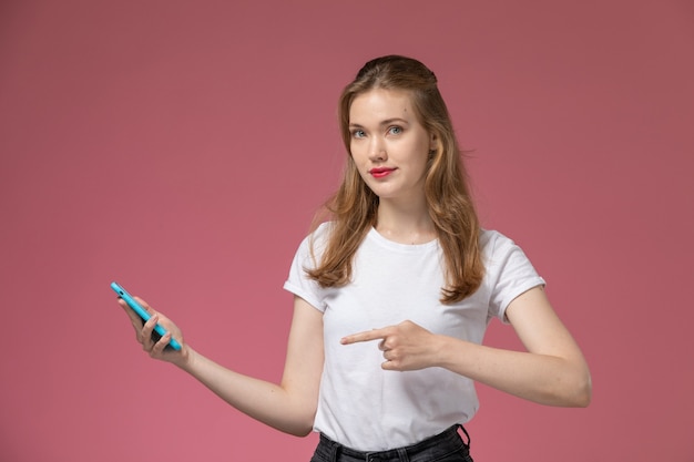 Front view young attractive female in white t-shirt posing holding smartphone on pink wall model female pose color photo female young