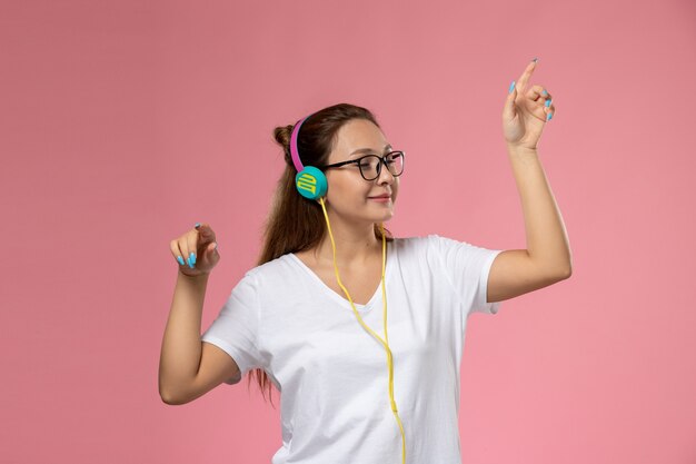 Front view young attractive female in white t-shirt posing listening to music with headphones and smile on the pink background