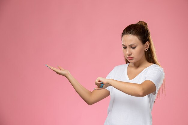 Front view young attractive female in white t-shirt posing and looking at her wrist on the pink background