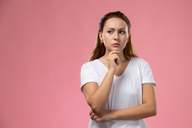 Front view young attractive female in white t-shirt posing and thinking on the pink background