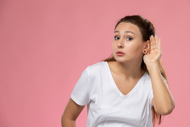 Front view young attractive female in white t-shirt posing and trying to hear out on the pink background