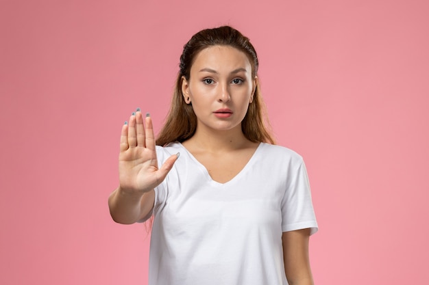 Front view young attractive female in white t-shirt posing with ban gesture on the pink background