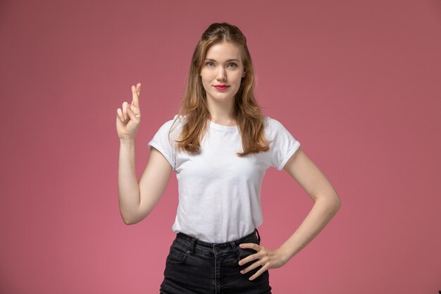 ピンクの壁のモデルの女性のポーズのカラー写真に交差した指でポーズをとる白いTシャツの正面図若い魅力的な女性