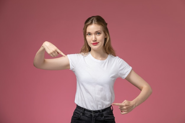 ピンクの壁のモデルの色の若い女性のジェスチャーでポーズをとる白いTシャツの正面図若い魅力的な女性