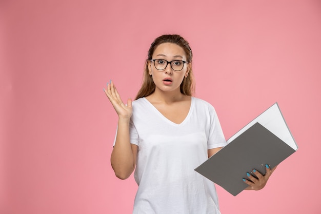 ピンクの背景に灰色のファイルでポーズをとって白いtシャツで正面の若い魅力的な女性