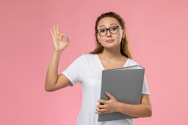 ピンクの背景に灰色のファイルでポーズをとって白いtシャツで正面の若い魅力的な女性