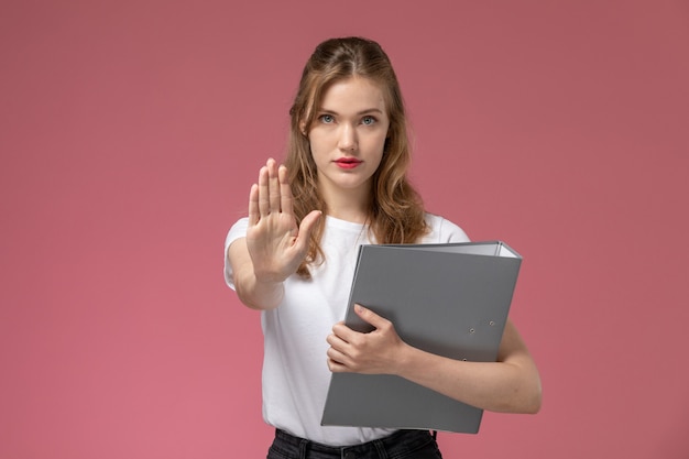 Front view young attractive female in white t-shirt posing with grey file showing stop sign on pink wall model color female young