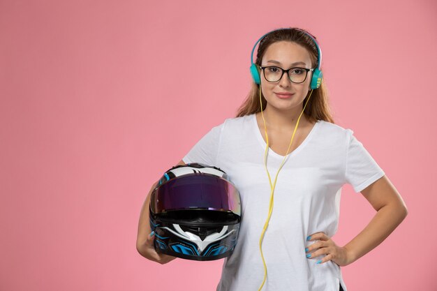 Front view young attractive female in white t-shirt posing with helmet listening to music on the pink background